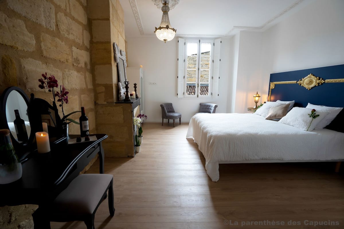 Un dormitorio con una cama, un escritorio con espejo, una silla y una ventana con vistas a edificios de piedra.
