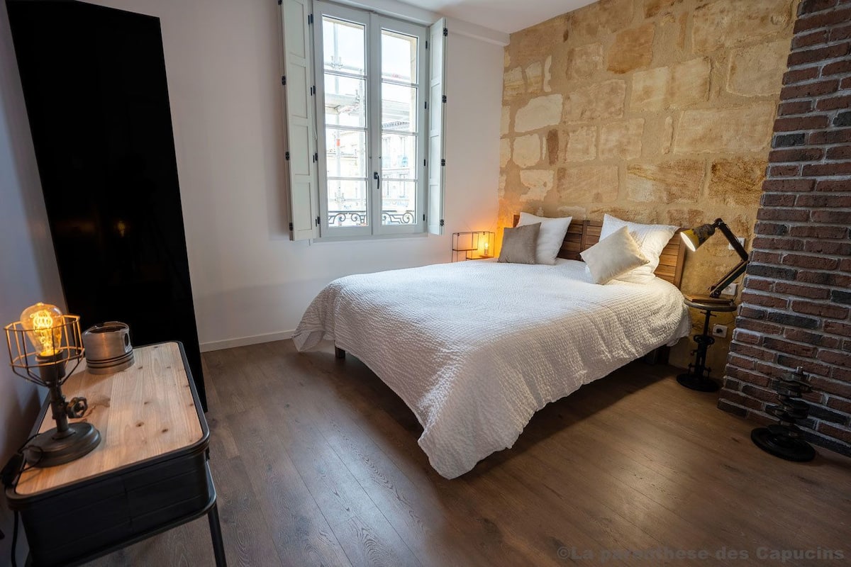 Un dormitorio con una cama grande, una mesita de noche de madera con una lámpara antigua, una pared de piedra y un ventanal con contraventanas blancas.