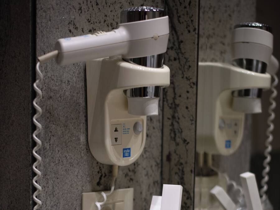 A bathroom with a hairdryer mounted on the wall, a mirror, and a countertop with a toothbrush holder, a glass of water, and a dental kit.