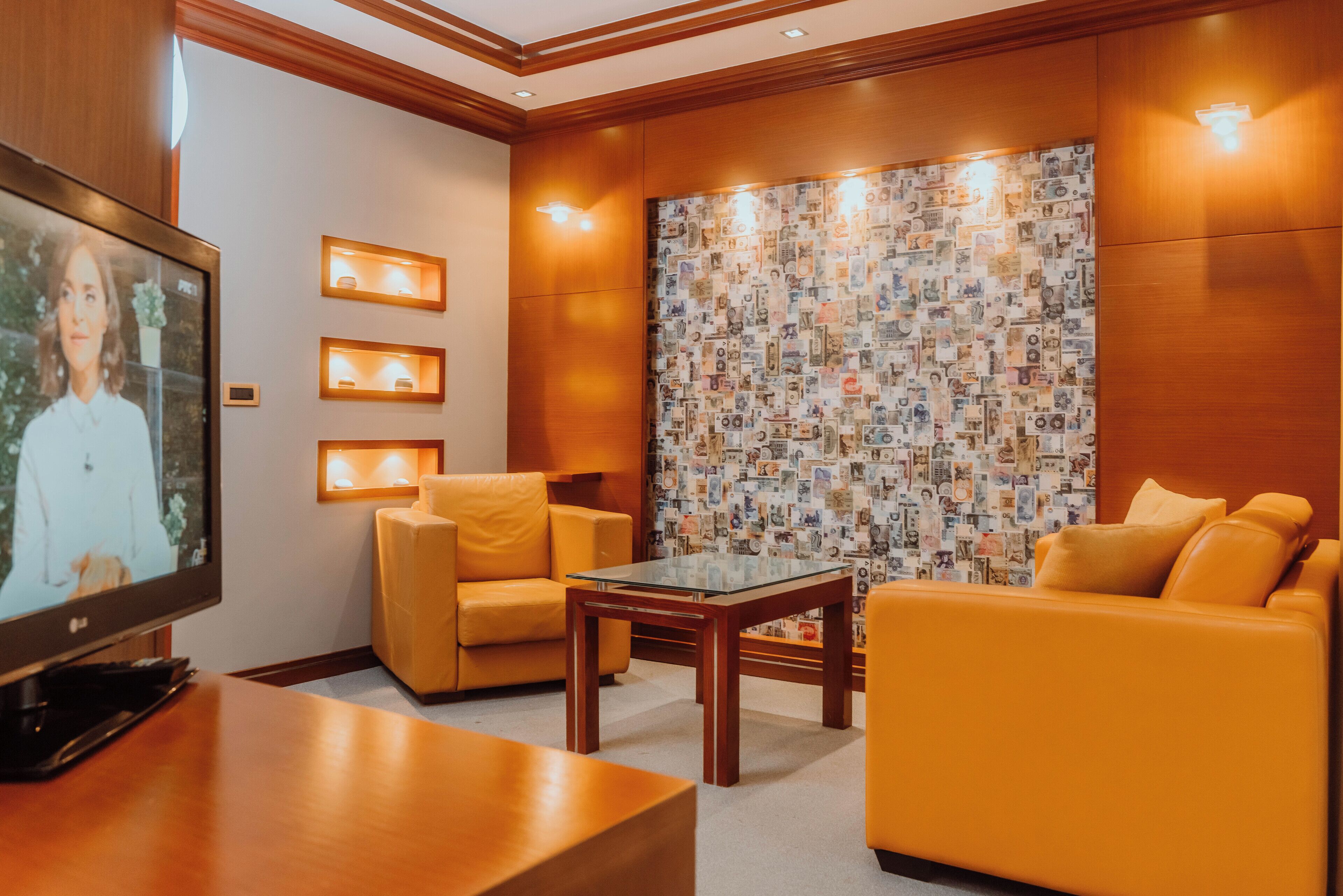 A room with a television, a sofa, a chair, and a wall with a currency-themed wallpaper.