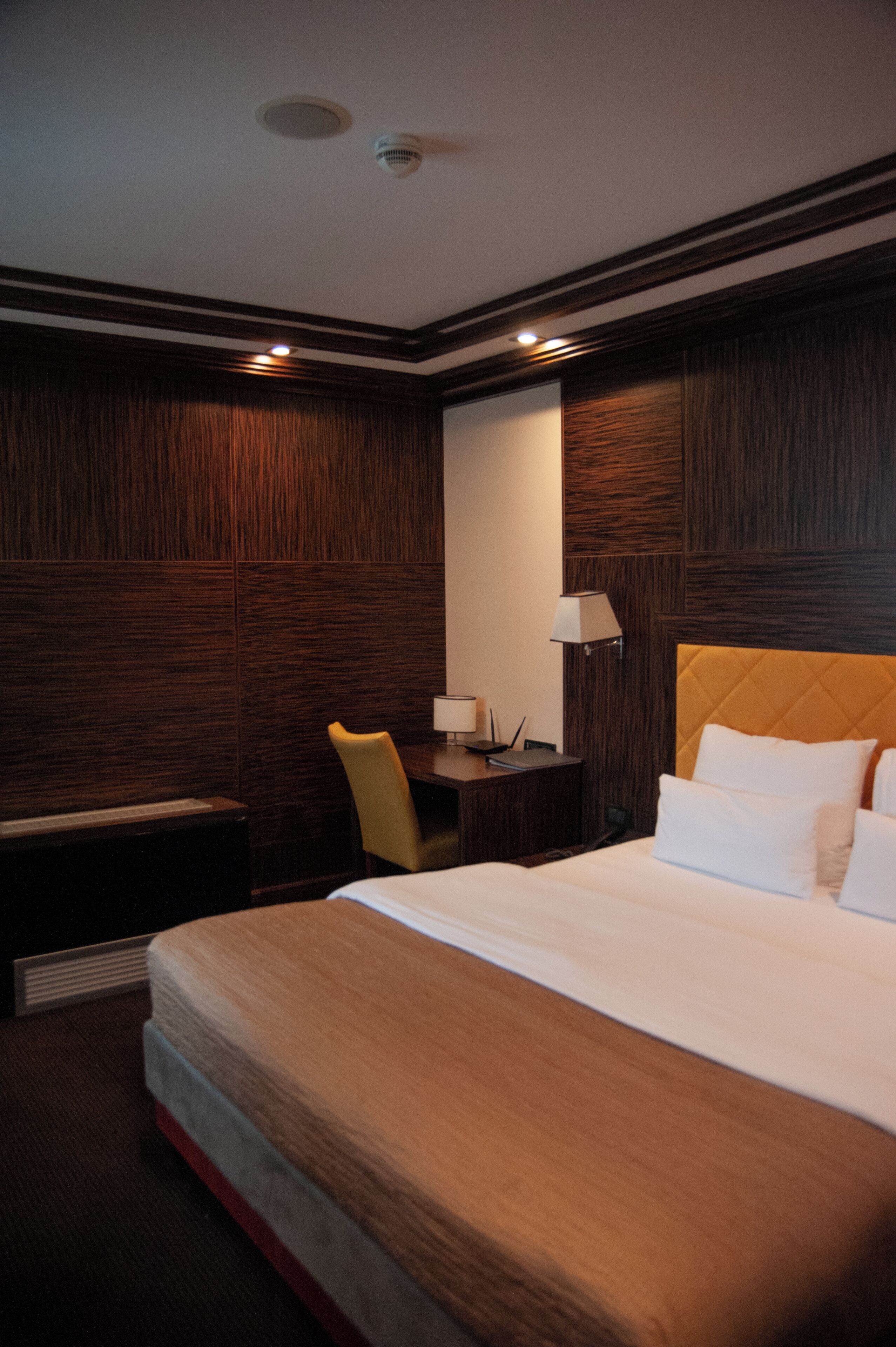 A hotel room with a large bed, a desk with a chair, a lamp, and a wall-mounted mirror.