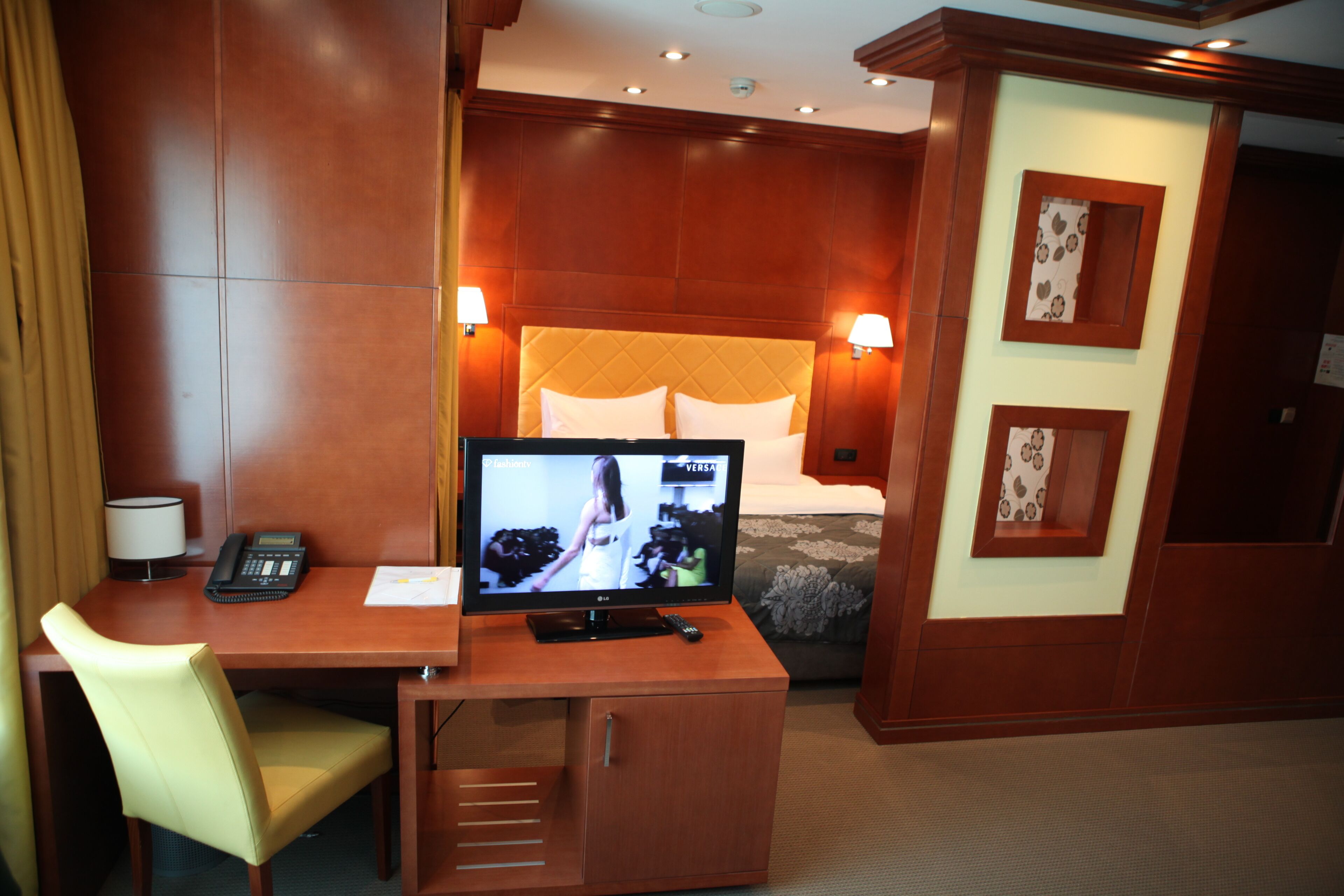 A hotel room with a wooden desk, a television, a bed with a quilted headboard, and two wall-mounted lamps.