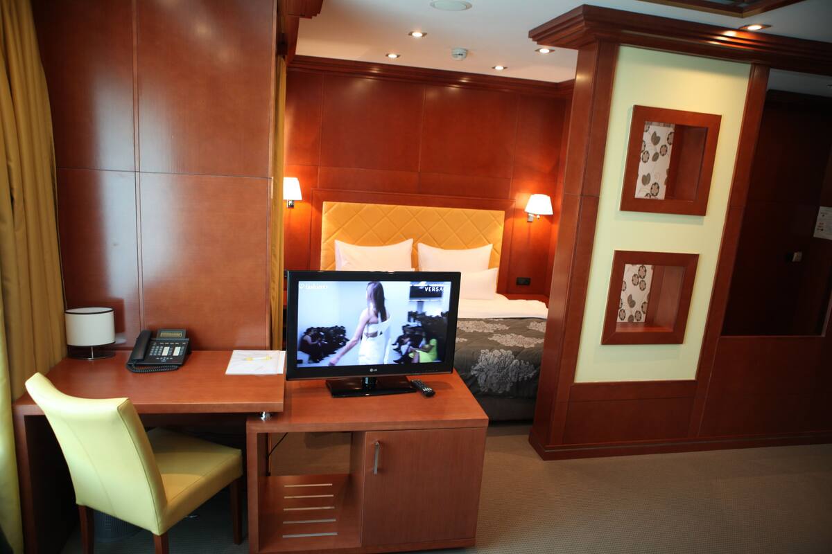 A hotel room with a wooden desk, a television, a bed with a quilted headboard, and two wall-mounted lamps.