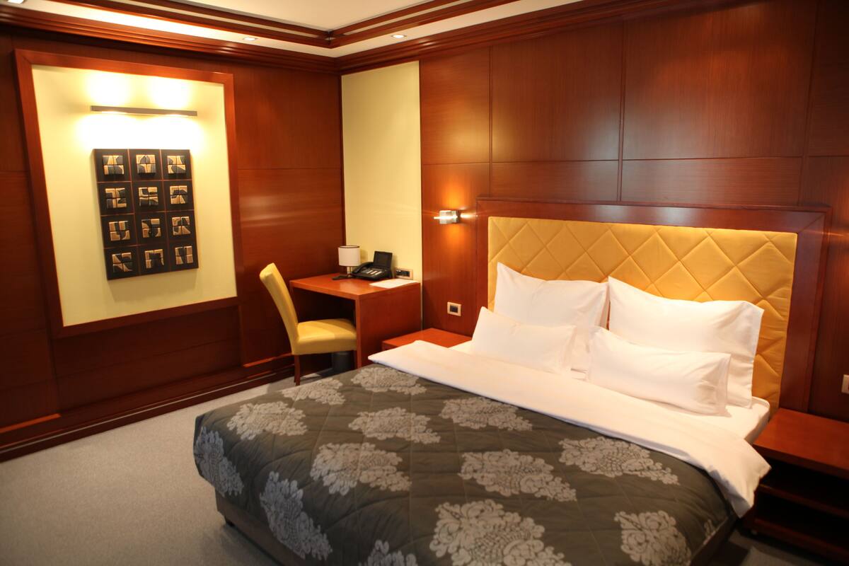 A hotel room with a large bed, a desk with a chair, and a wall-mounted light fixture.