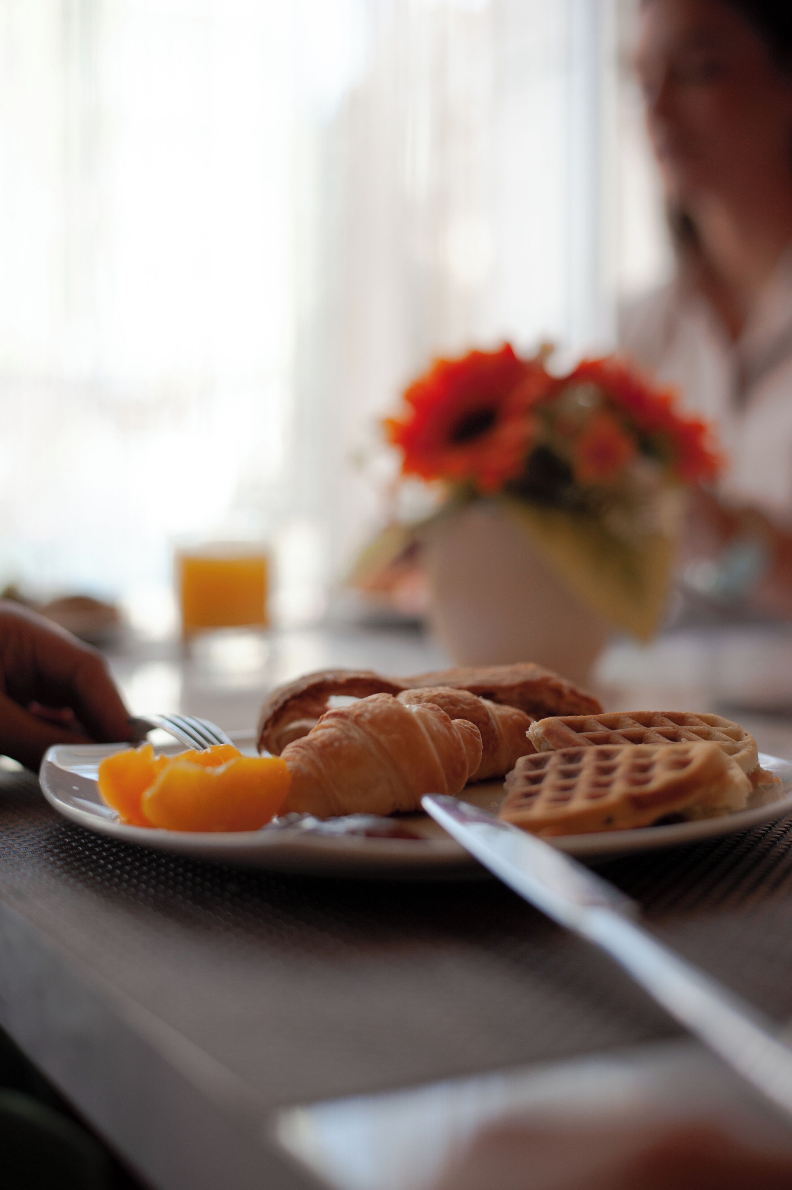 A breakfast setting with a plate of food, a flower vase, and a blurred person.