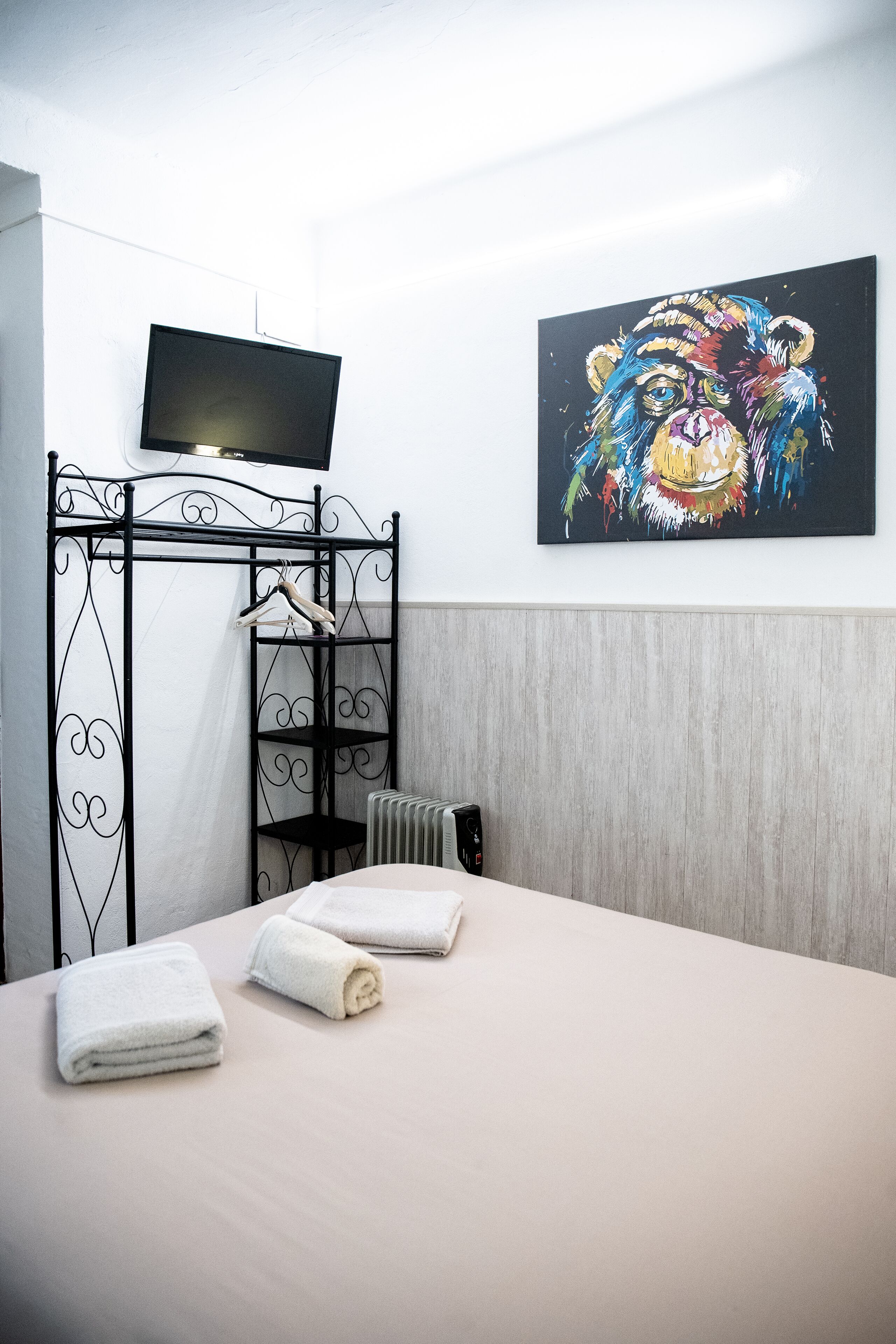 A hotel room with a bed, a TV mounted on the wall, a colorful artwork, and a black metal wardrobe.
