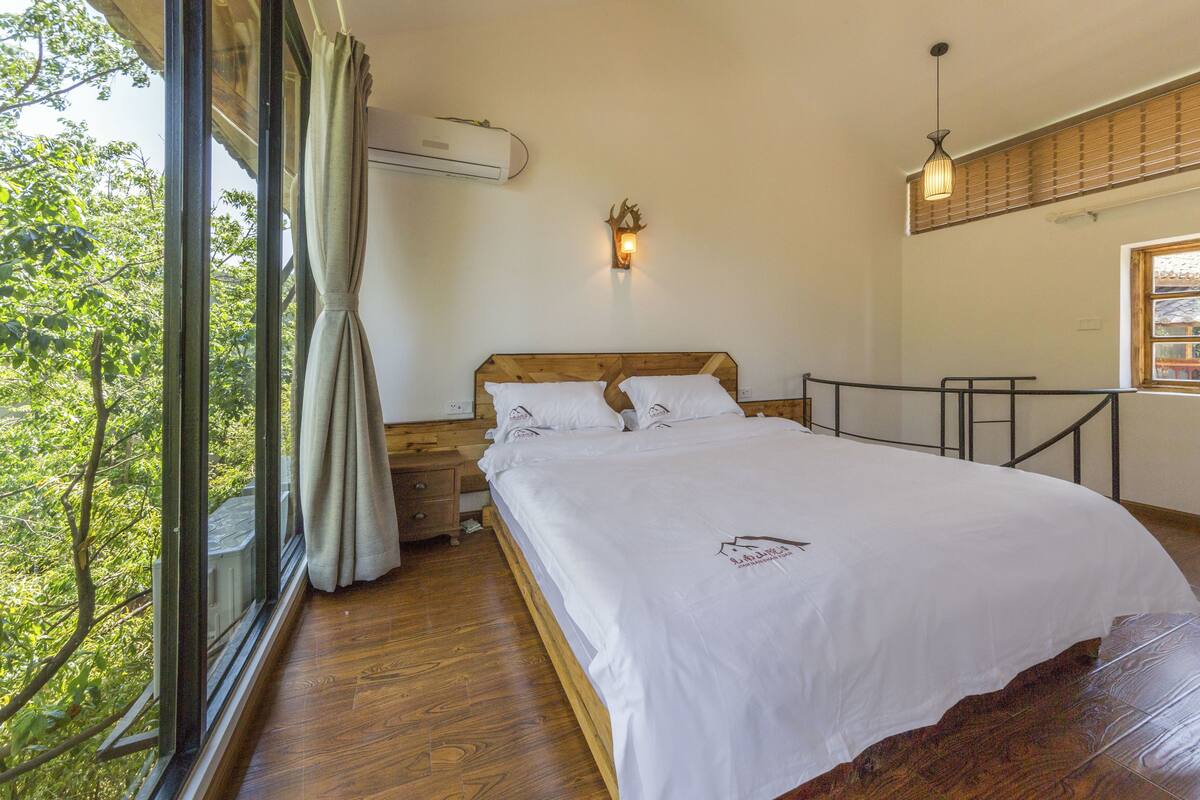 A bedroom with a wooden bed, white bedding, a nightstand, a wall-mounted light, and a window with curtains.