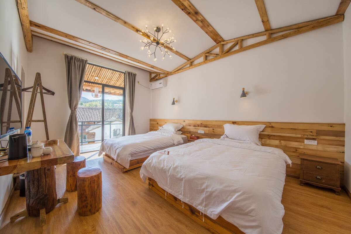 A room with two beds, wooden beams, a chandelier, a wooden desk, and a window with curtains.
