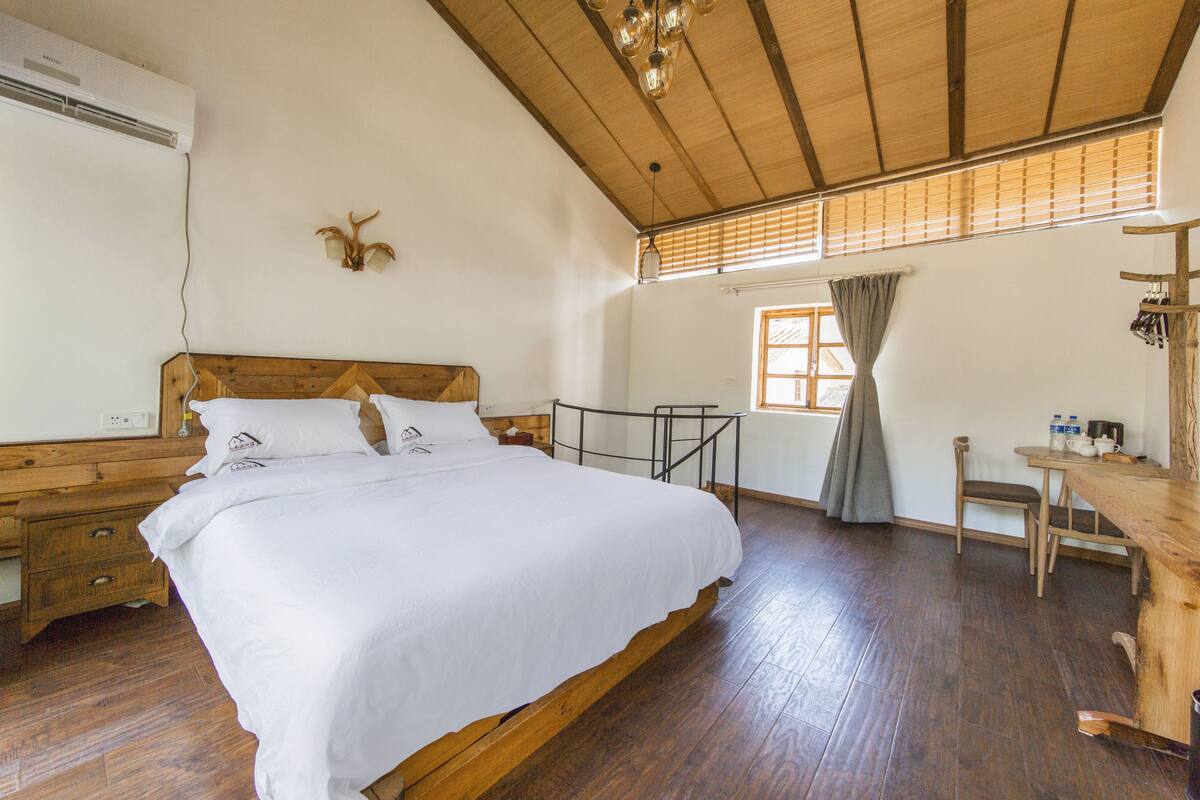 A room with a wooden bed, a desk with a chair, a window with curtains, and a wooden ceiling.