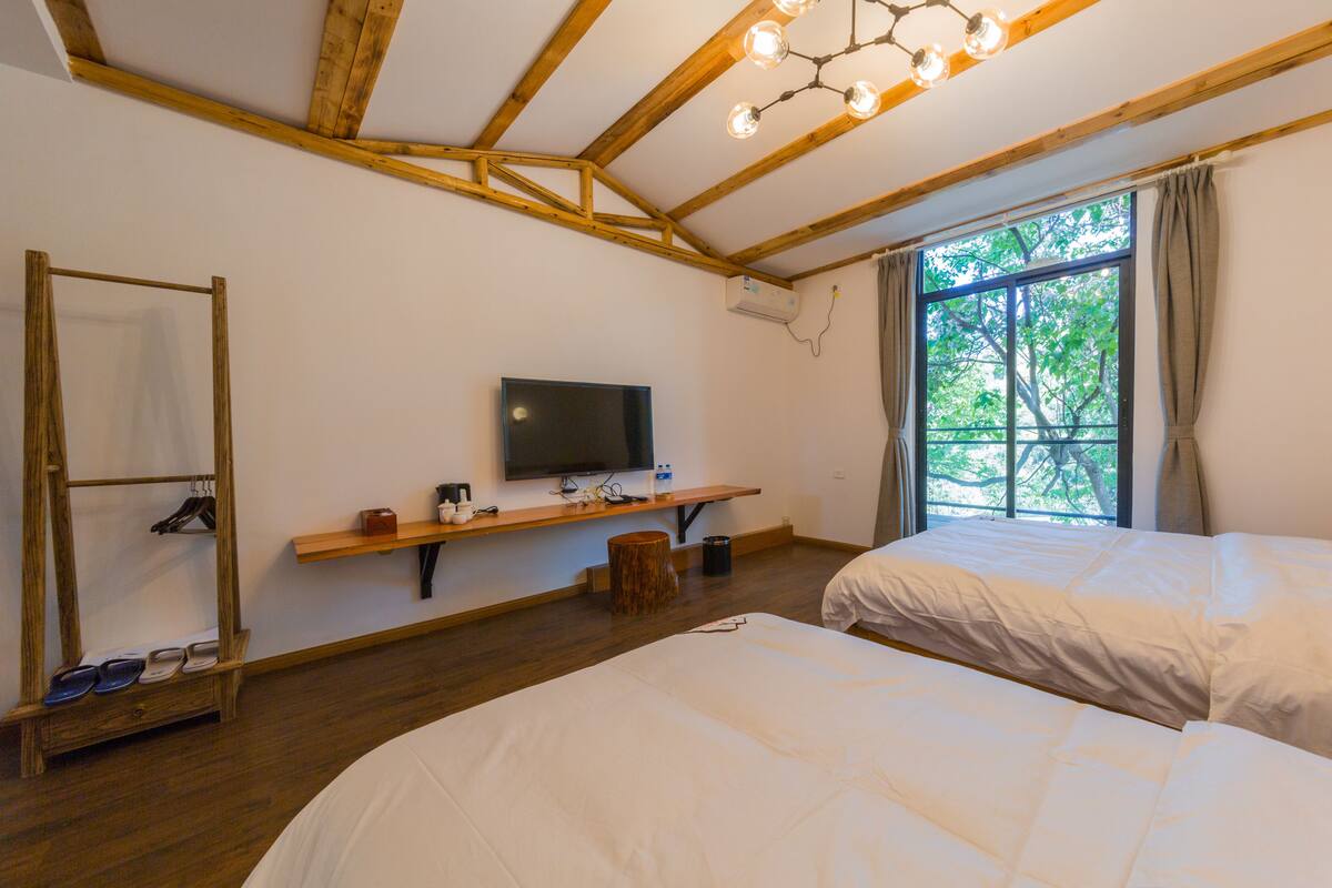 A room with two beds, a wooden ceiling, a mounted TV, and a window with curtains.