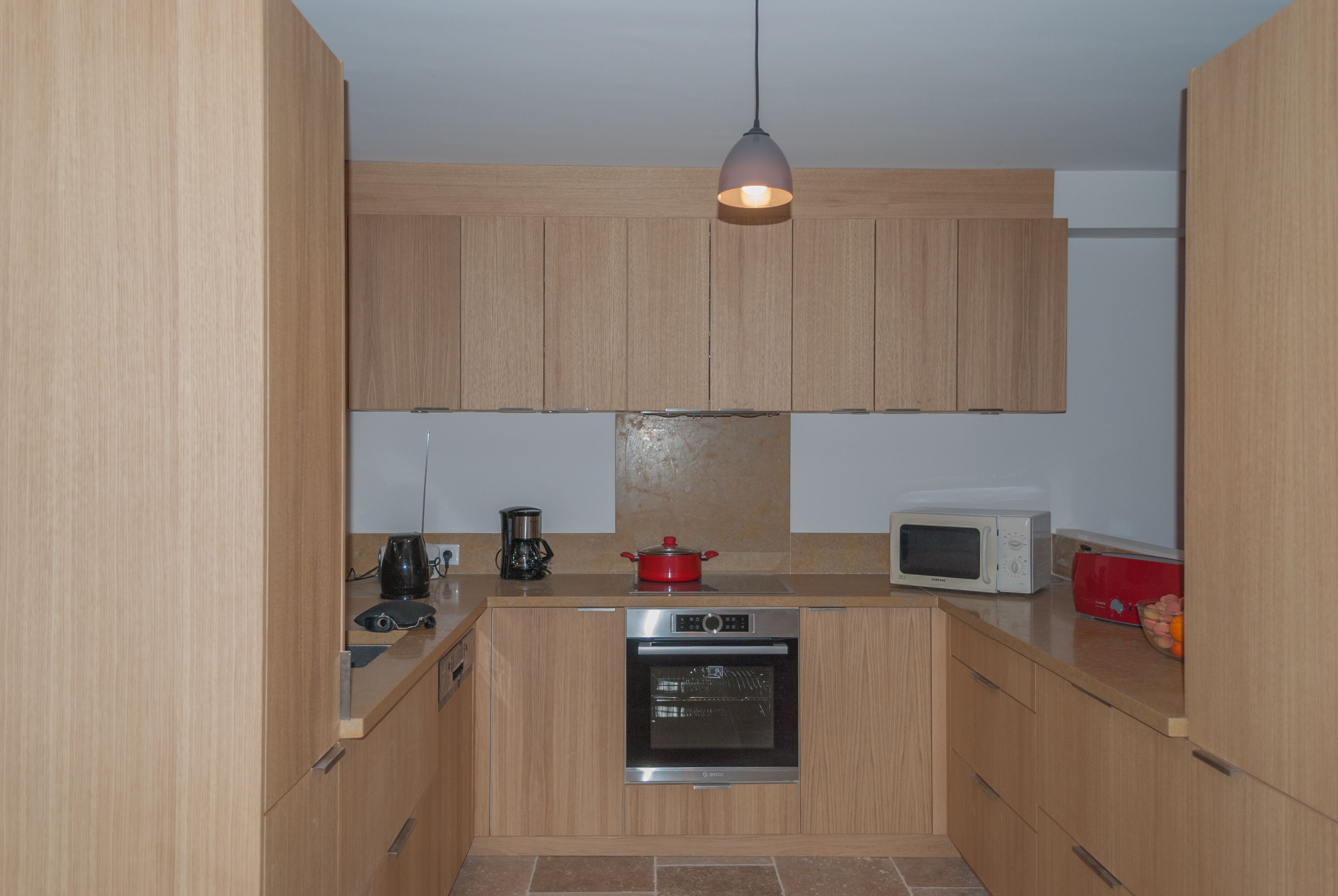 Une cuisine compacte avec des meubles en bois, un four encastré, un four à micro-ondes et un luminaire suspendu.
