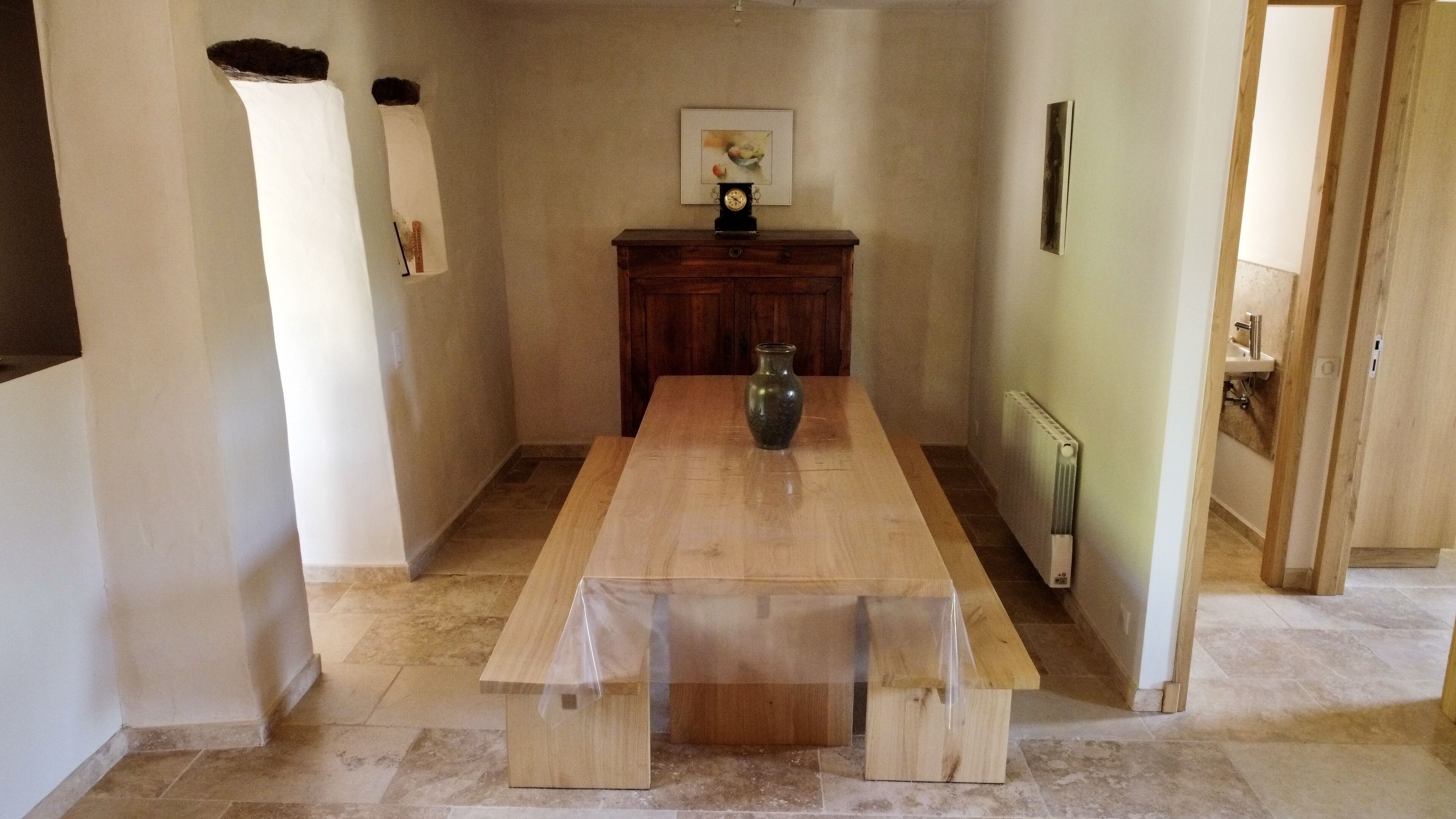 A dining area with a wooden table and benches, a vase, and a clock on a cabinet.