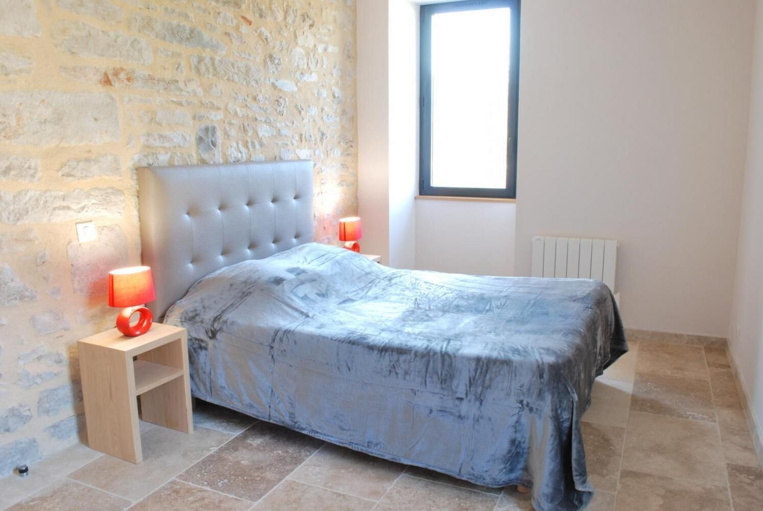 Un dormitorio con pared de piedra, una cama con cubierta gris, dos lámparas de noche y un ventanal.