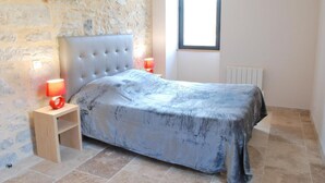 A bedroom with a stone wall, a bed with a grey cover, two bedside lamps, and a window. - Gite Le Roucayral– Superb view over St Cirq Lapopie – 2 air-conditioned bedrooms (Tour de Faure)