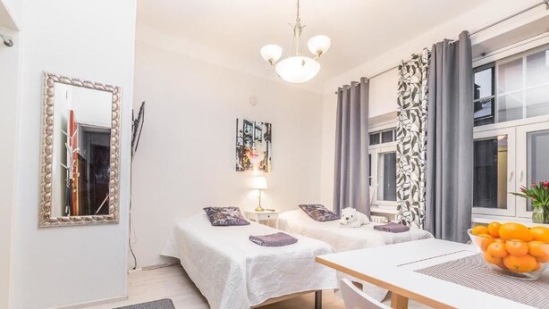 A hotel room with two beds, a dining table, a mirror, a lamp, and a window with curtains. - Go Happy Home Apartment Mikonkatu 18 (Helsinki)