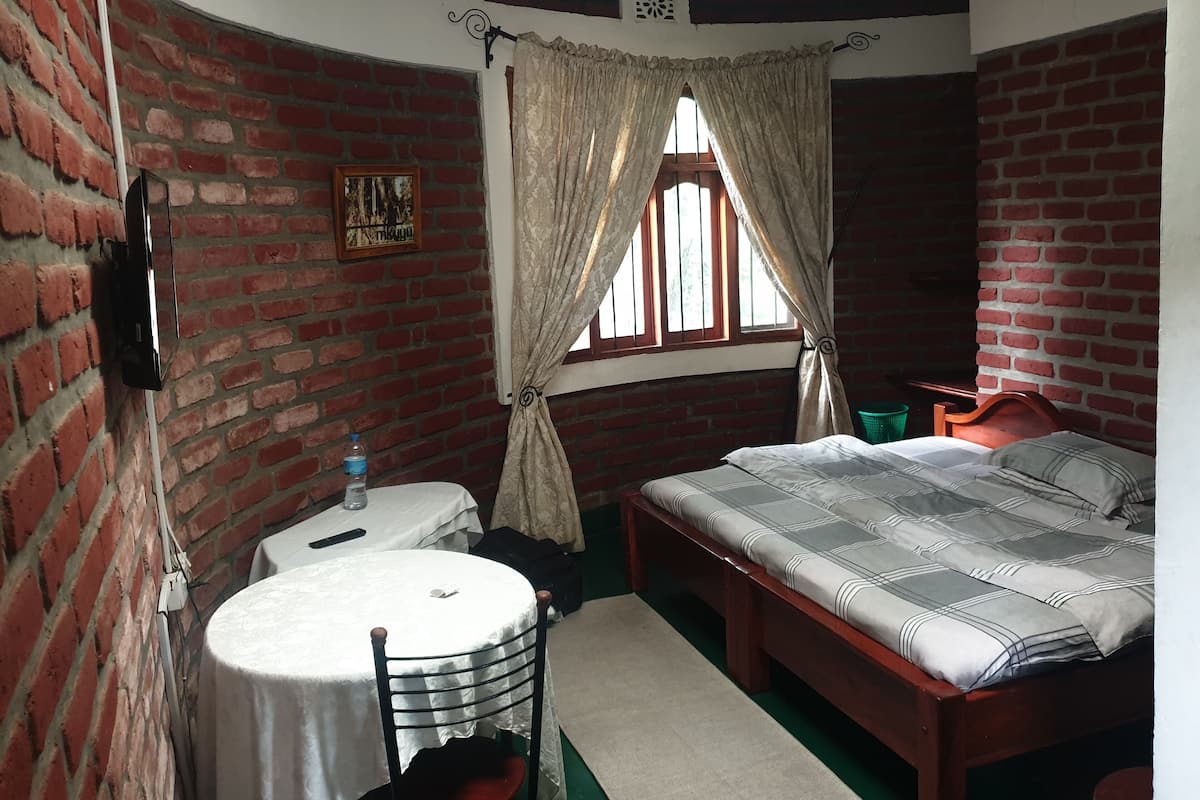 A room with a bed, a table, and a chair. There is a window with curtains, a brick wall, and a picture on the wall.