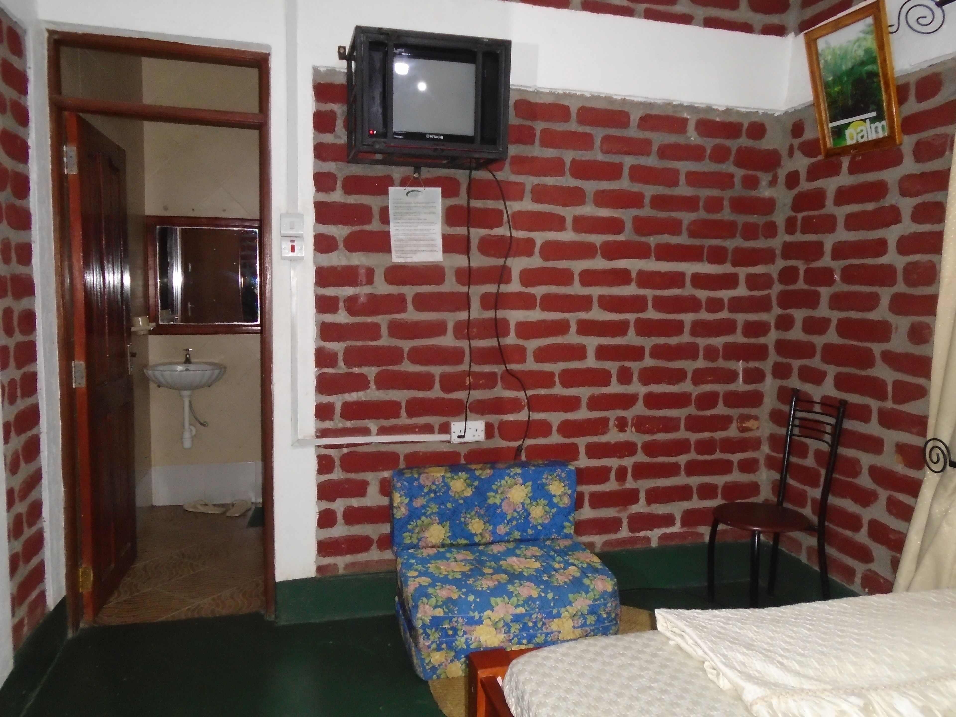 Una habitación con pared de ladrillos, televisor sobre un lavamanos, silla con estampado floral y cama con cubrecama blanco.