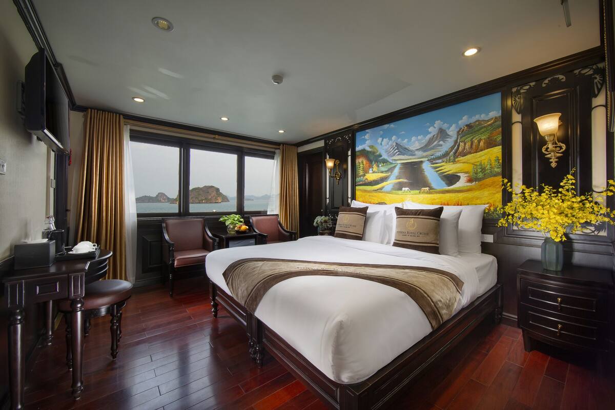 A large bed with a view of the mountains through the window, a desk with a chair, and a vase of yellow flowers.