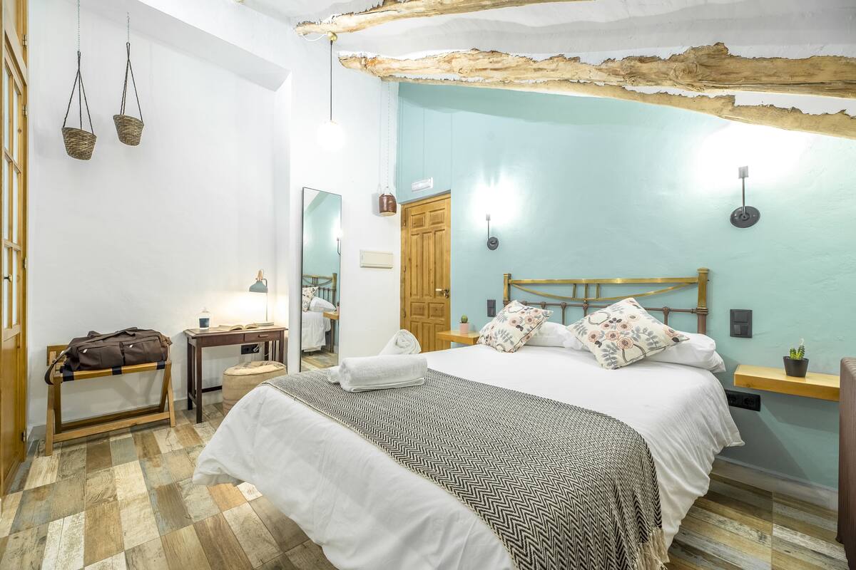 A bedroom with a bed, bedside tables, a wooden headboard, a hanging basket, a mirror, and a wooden door.