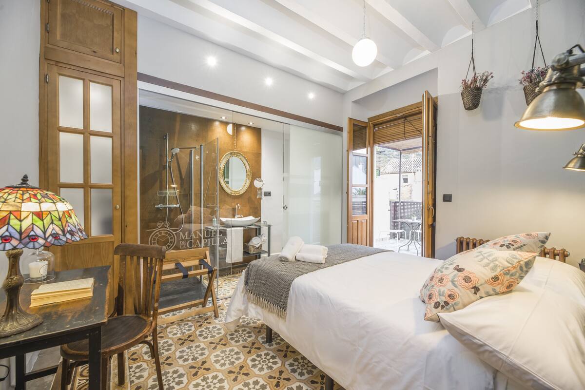 A hotel room with a bed, a desk with a lamp, a chair, a bathroom with a mirror, and a view of the outside.
