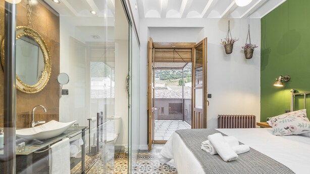 A modern bathroom with a glass-enclosed sink, a large mirror, and a shower. The room features a patterned floor, a bed with neatly folded towels, and a view through an open door to an outdoor area. - Manuel de La Capilla (Cambil)
