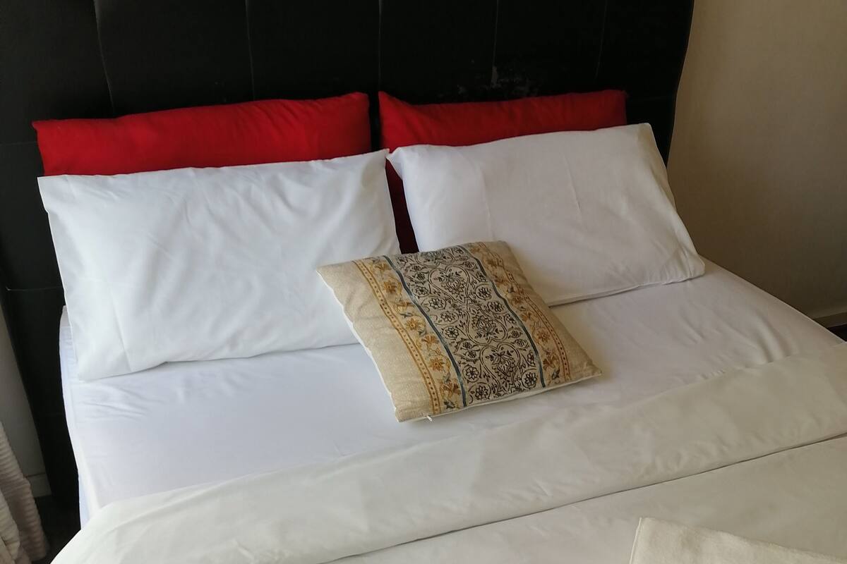 Un lit bien fait, avec une literie blanche, des coussins rouges et beiges, et un coussin décoratif orné d’un motif floral.