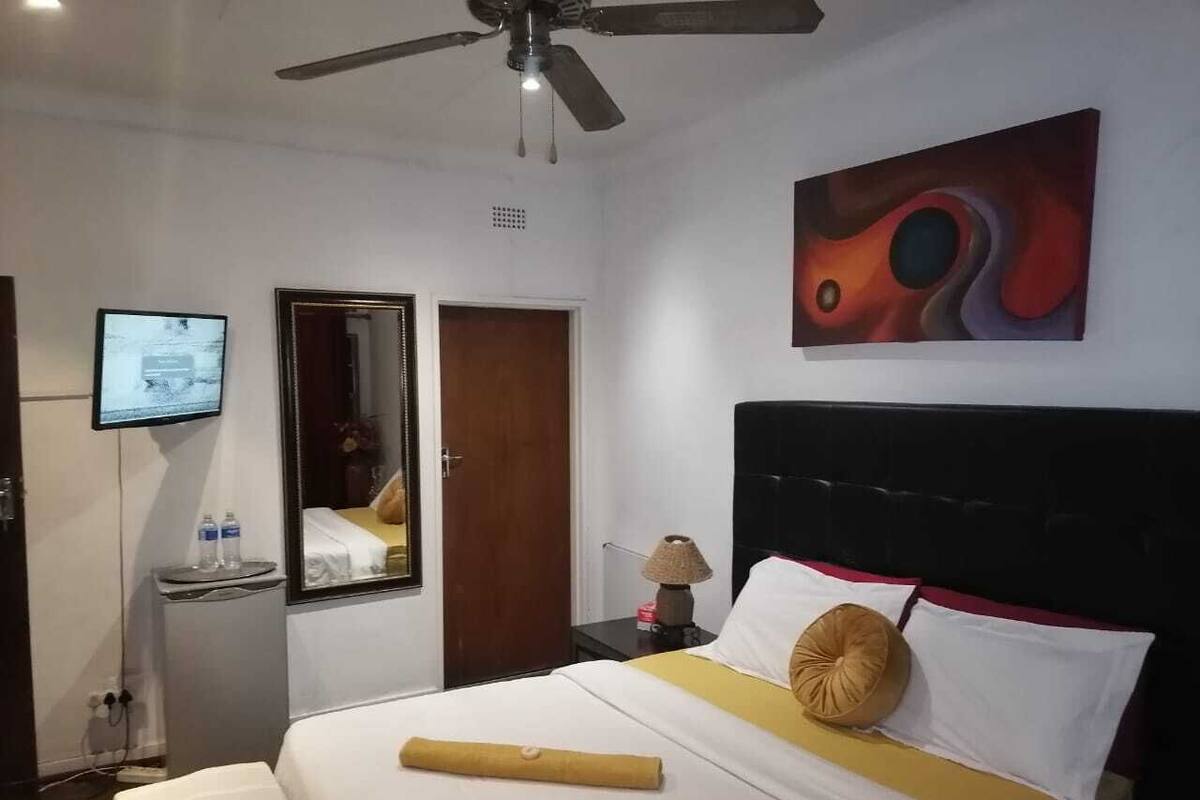 Une chambre d’hôtel avec un lit, un ventilateur de plafond, une télévision, un miroir, une table de chevet avec une lampe et un tableau au mur.