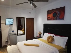 Une chambre d’hôtel avec un lit, un ventilateur de plafond, une télévision, un miroir, une table de chevet avec une lampe et un tableau au mur.