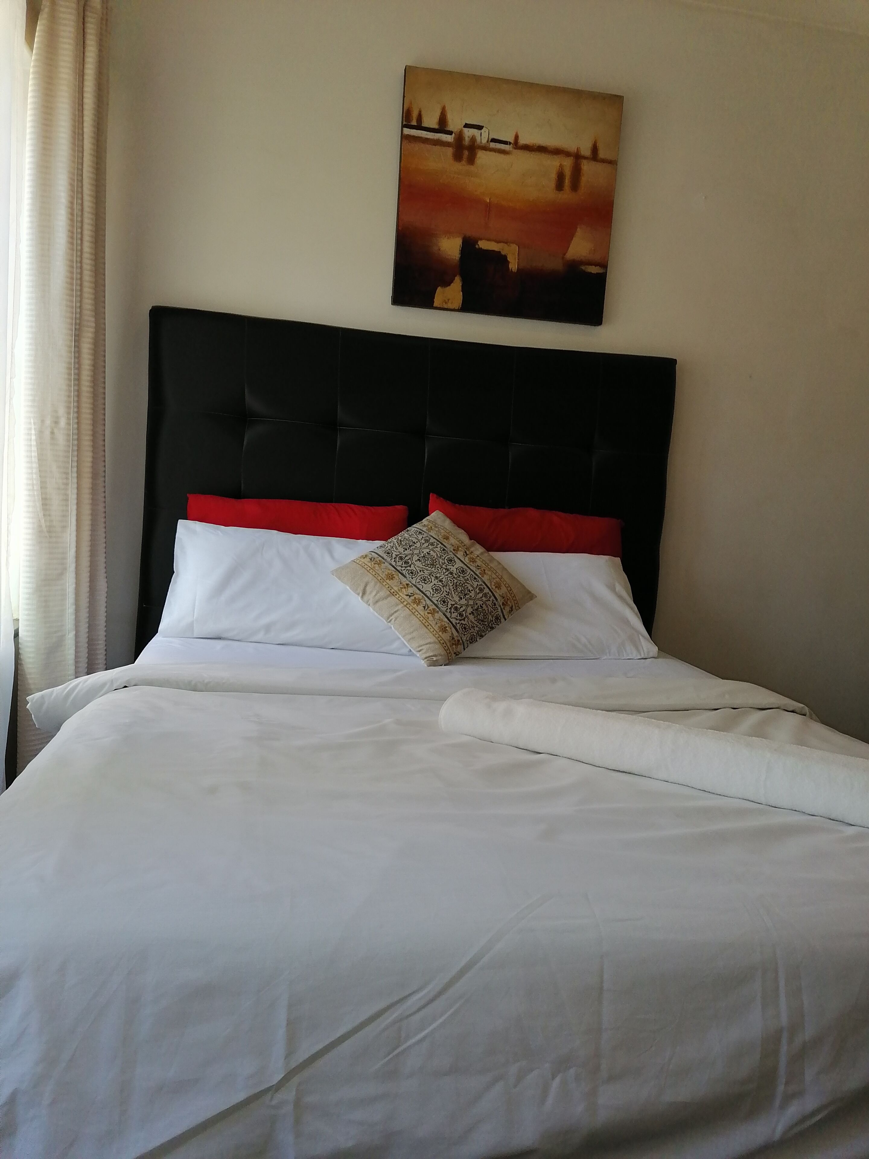Een net opgemaakt bed met wit beddengoed en een rode sierkussen, een zwart bekleed hoofdbord, en een schilderij aan de muur erboven.