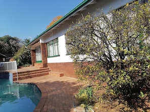 Une maison avec une piscine, un chemin en briques et un jardin agrémenté d’arbres et d’arbustes.