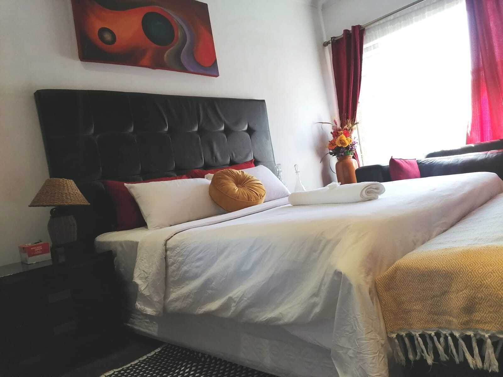 Una camera d'albergo con testiera trapuntata, un letto con lenzuola bianche, un cuscino rosso e bianco, una coperta gialla, una lampada e un quadro appeso al muro.
