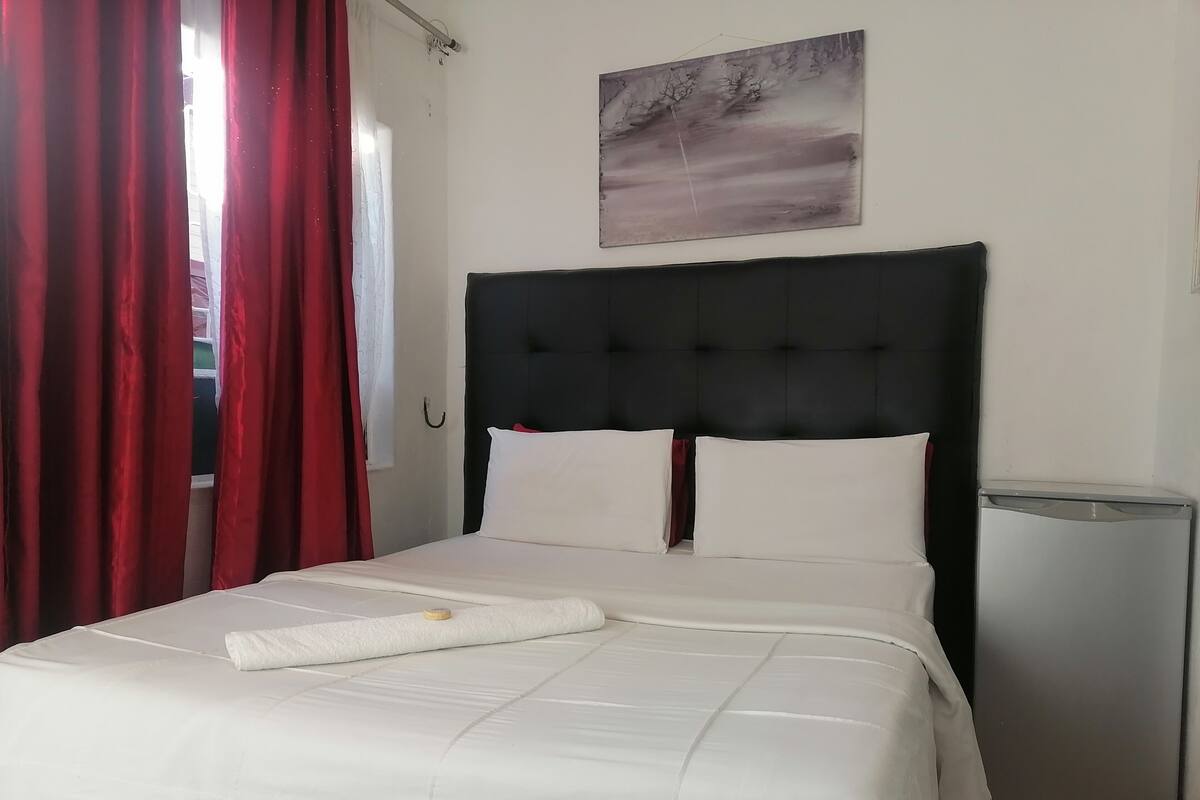 Un lit bien fait, avec du linge de lit blanc et une serviette pliée, une tête de lit rembourrée de couleur sombre, une petite table de chevet et un tableau accroché au mur.