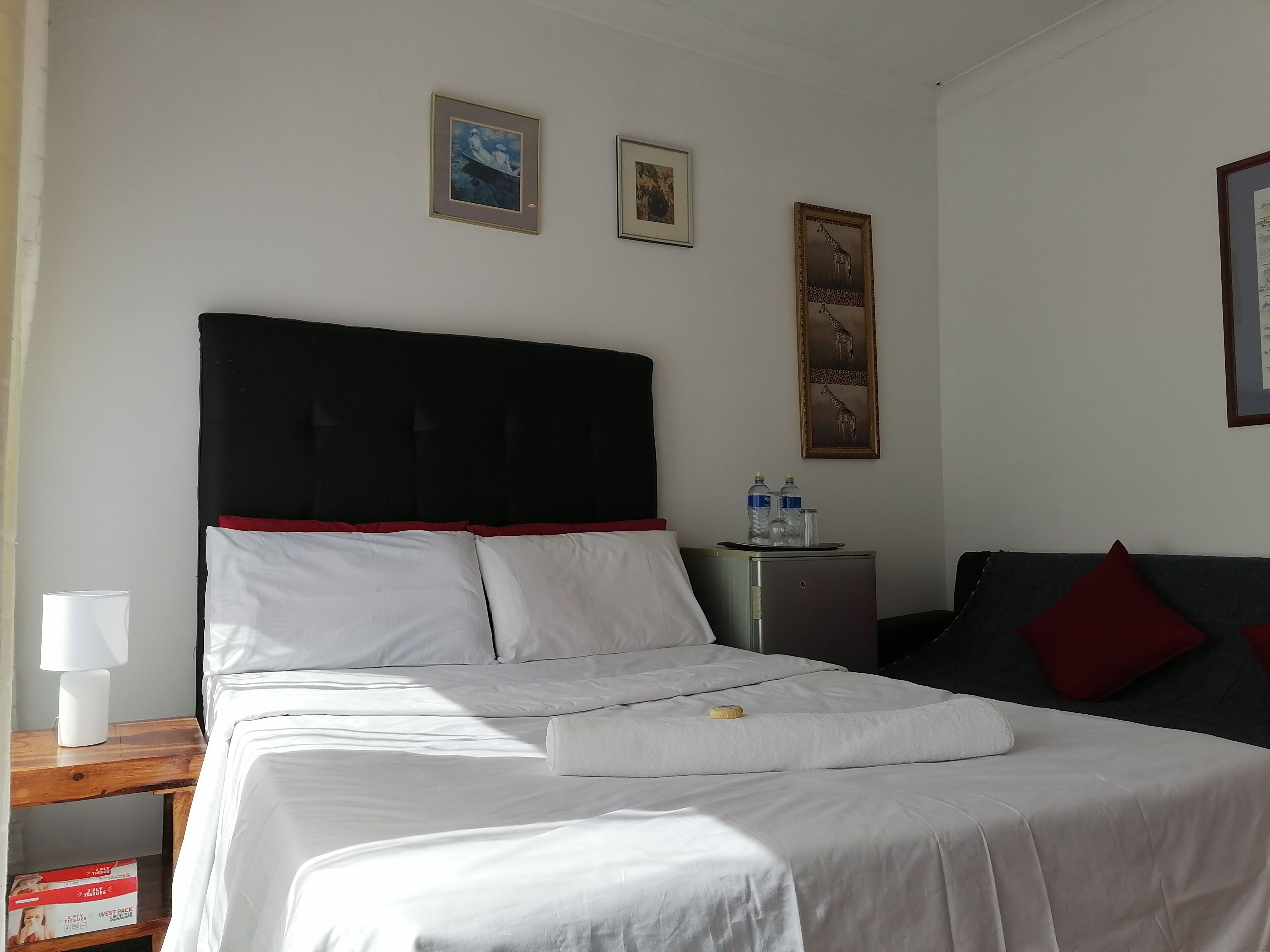 A hotel room with a bed, a nightstand, a lamp, a small table with water bottles, and framed pictures on the wall.
