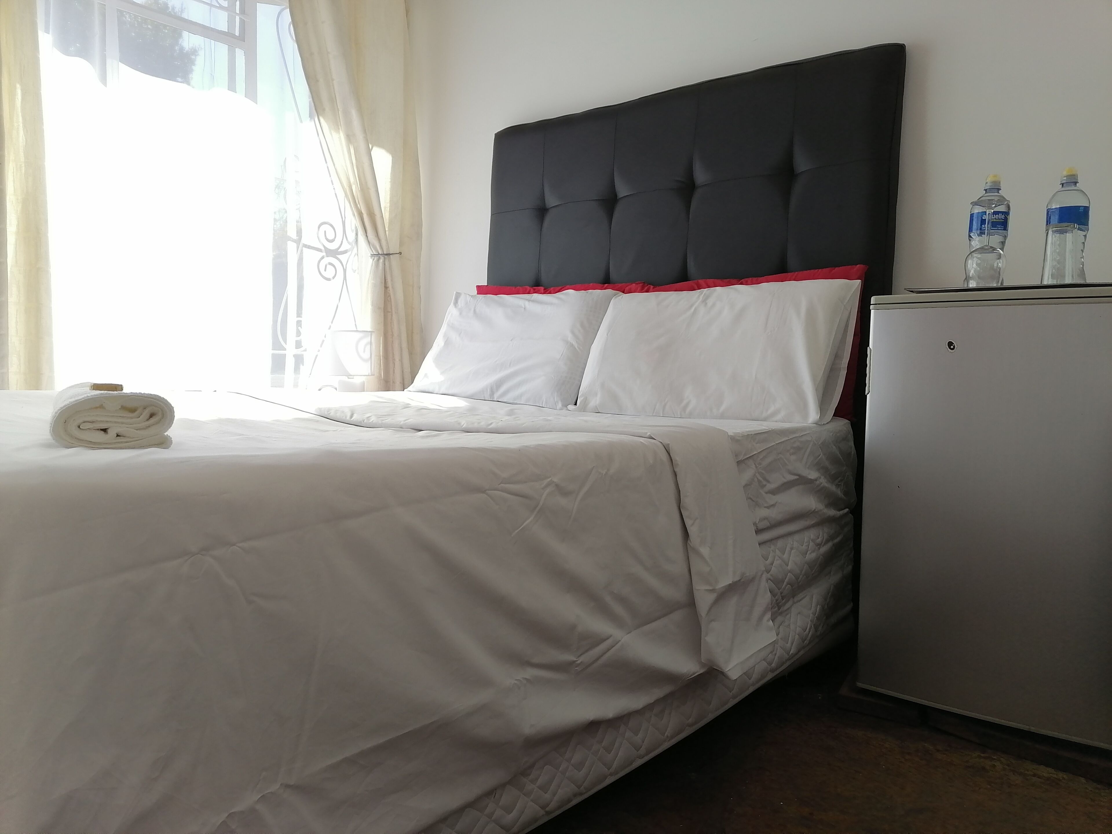 A neatly made bed with white bedding and a black headboard with red trim. There are two water bottles on a small white cabinet beside the bed.