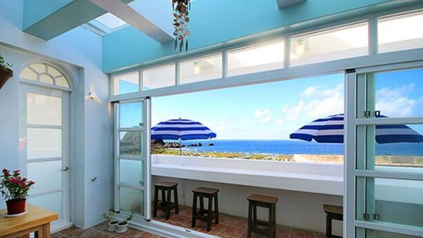 Quarto com uma grande janela com vista para a praia, uma porta e uma pequena mesa com uma planta em vaso.