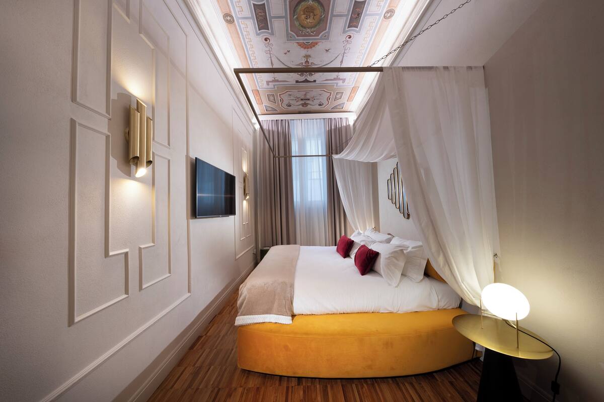 A hotel room with a curved bed, a flat-screen TV, a bedside table with a lamp, and a decorative ceiling.