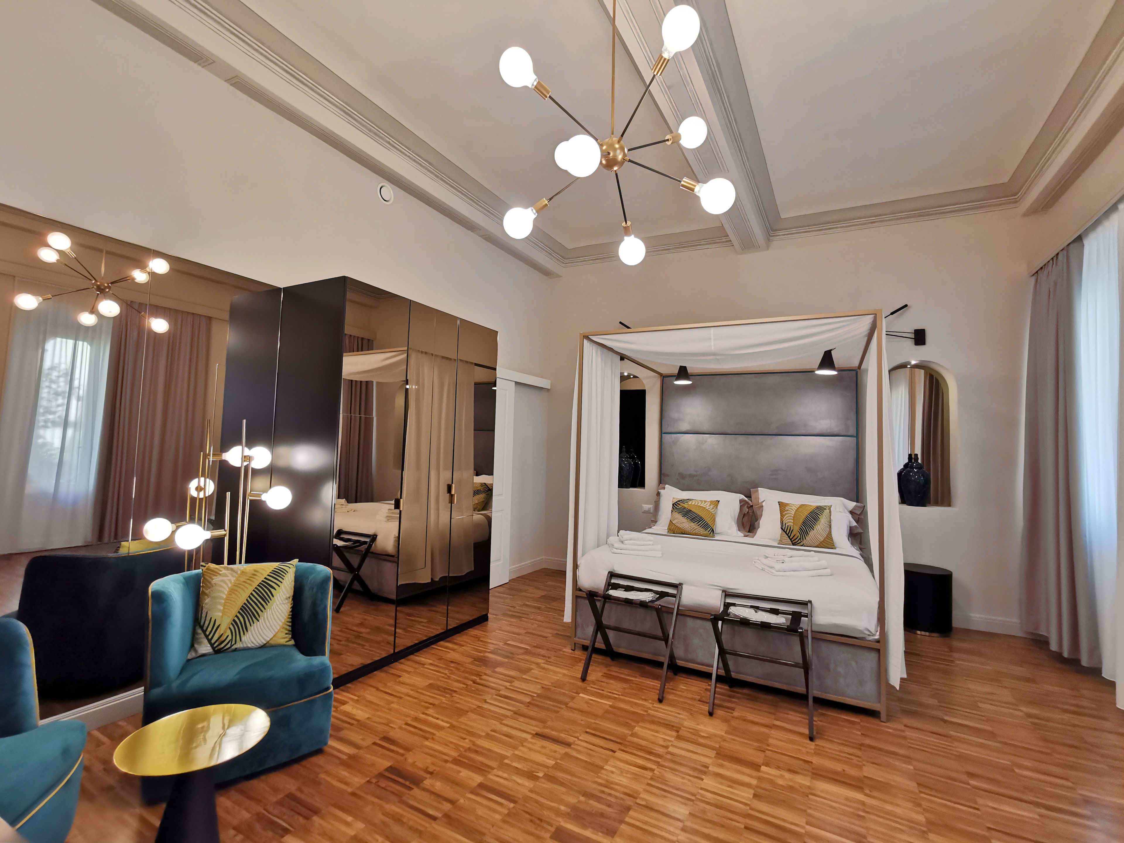 A modern hotel room with a large bed, a vanity with mirrors, a blue armchair, and a wooden floor.