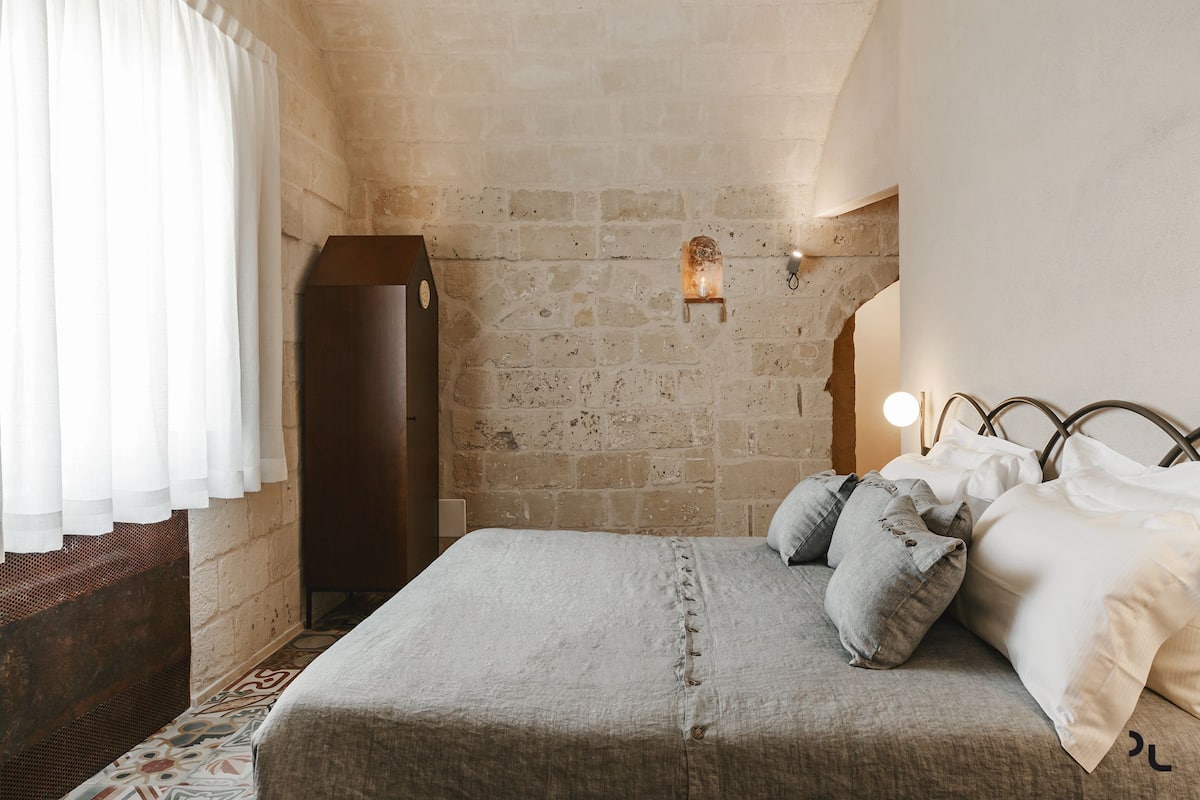 A bedroom with a stone wall, a bed with grey bedding, and a nightstand.