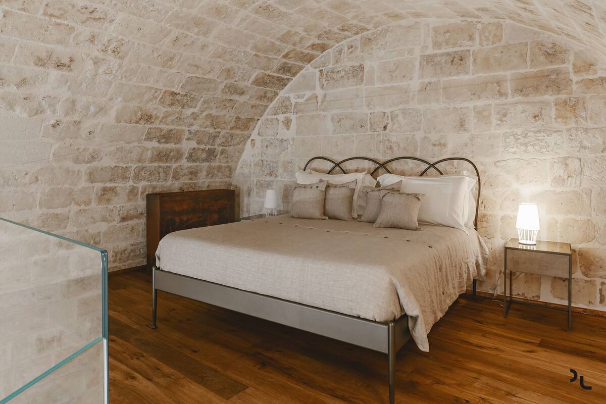 A bedroom with a stone wall, a metal bed frame, a wooden floor, and a bedside table with a lamp.