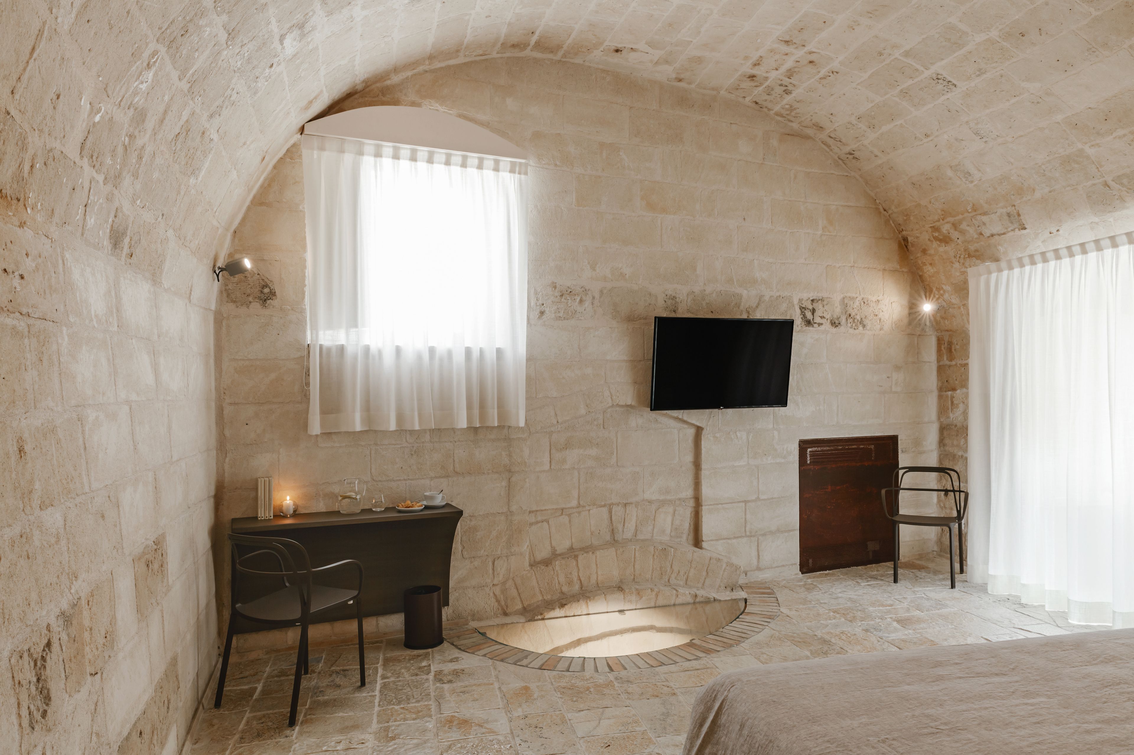 A stone-walled room with a fireplace, a desk with a chair, a television mounted on the wall, and a window with curtains.