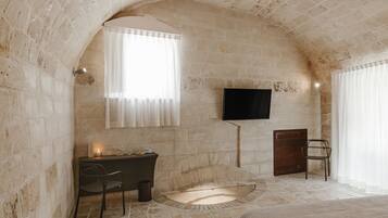 A stone-walled room with a fireplace, a desk with a chair, a television mounted on the wall, and a window with curtains.