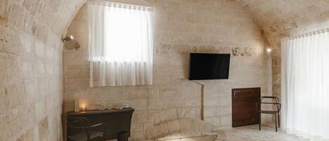A stone-walled room with a fireplace, a desk with a chair, a television mounted on the wall, and a window with curtains.