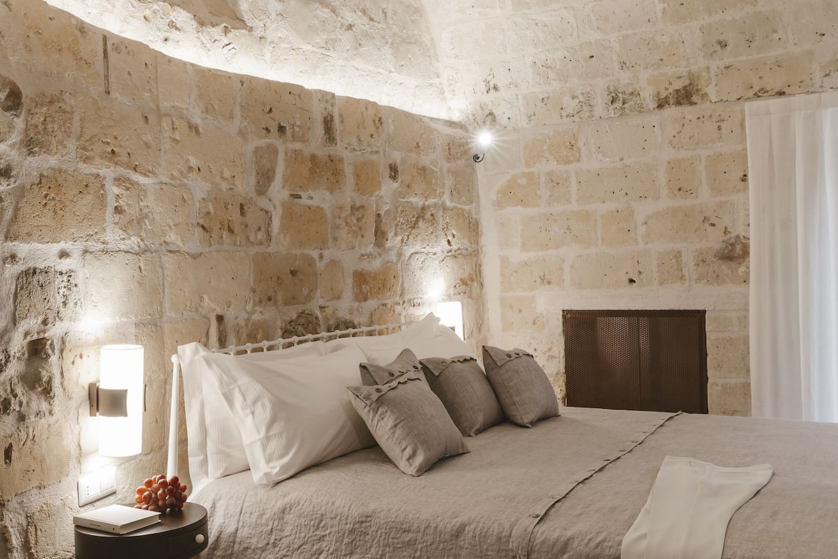 A bedroom with a stone wall, a bed with a headboard, a bedside table with a lamp, and patterned floor tiles.