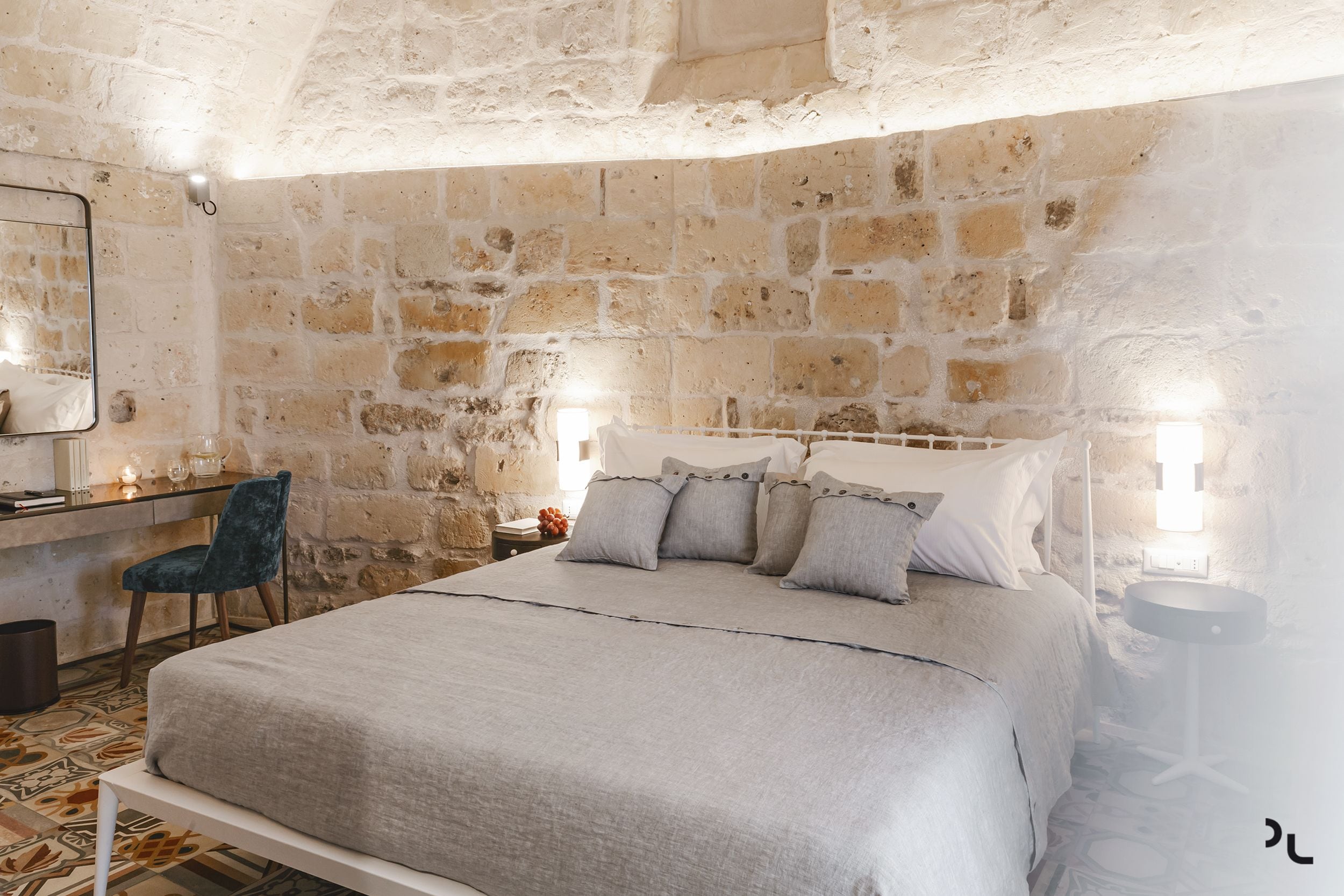 A bedroom with a stone wall, a bed with grey bedding, a desk with a chair, and bedside lamps.