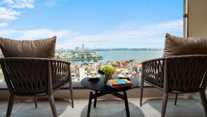 Un balcon avec des fauteuils en osier, une petite table ornée d’une plante et d’un livre, et une vue sur un paysage urbain qui s’étend de l’autre côté d’une étendue d’eau.
