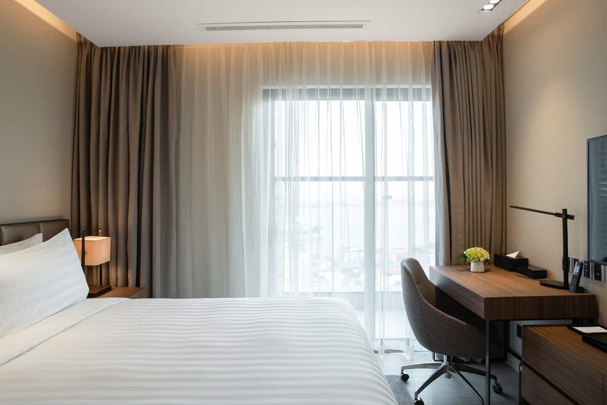 Ett hotellrum med en stor säng, ett skrivbord med en stol, en lampa och utsikt över staden genom tunna gardiner.
