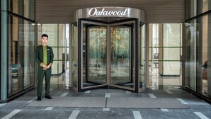 Une porte tournante surmontée d’une enseigne indiquant « Oakwood », une personne debout devant, et la façade d’un immeuble moderne avec des fenêtres en verre.
