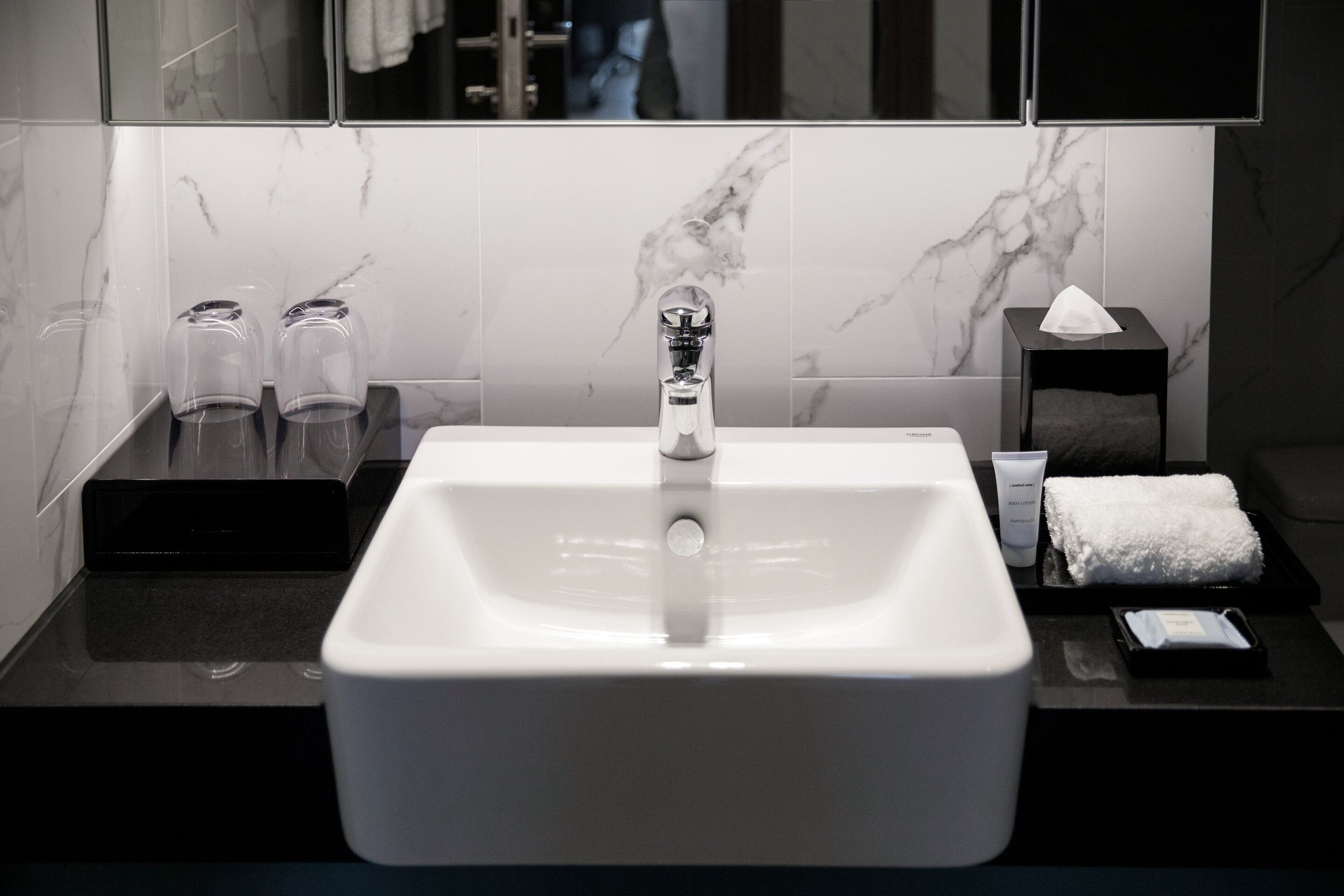 Une salle de bain moderne avec un grand lavabo blanc, un miroir et un pare-baignoire en marbre. On y trouve deux pots en verre sur le comptoir, une serviette et un petit plateau contenant des articles de toilette.