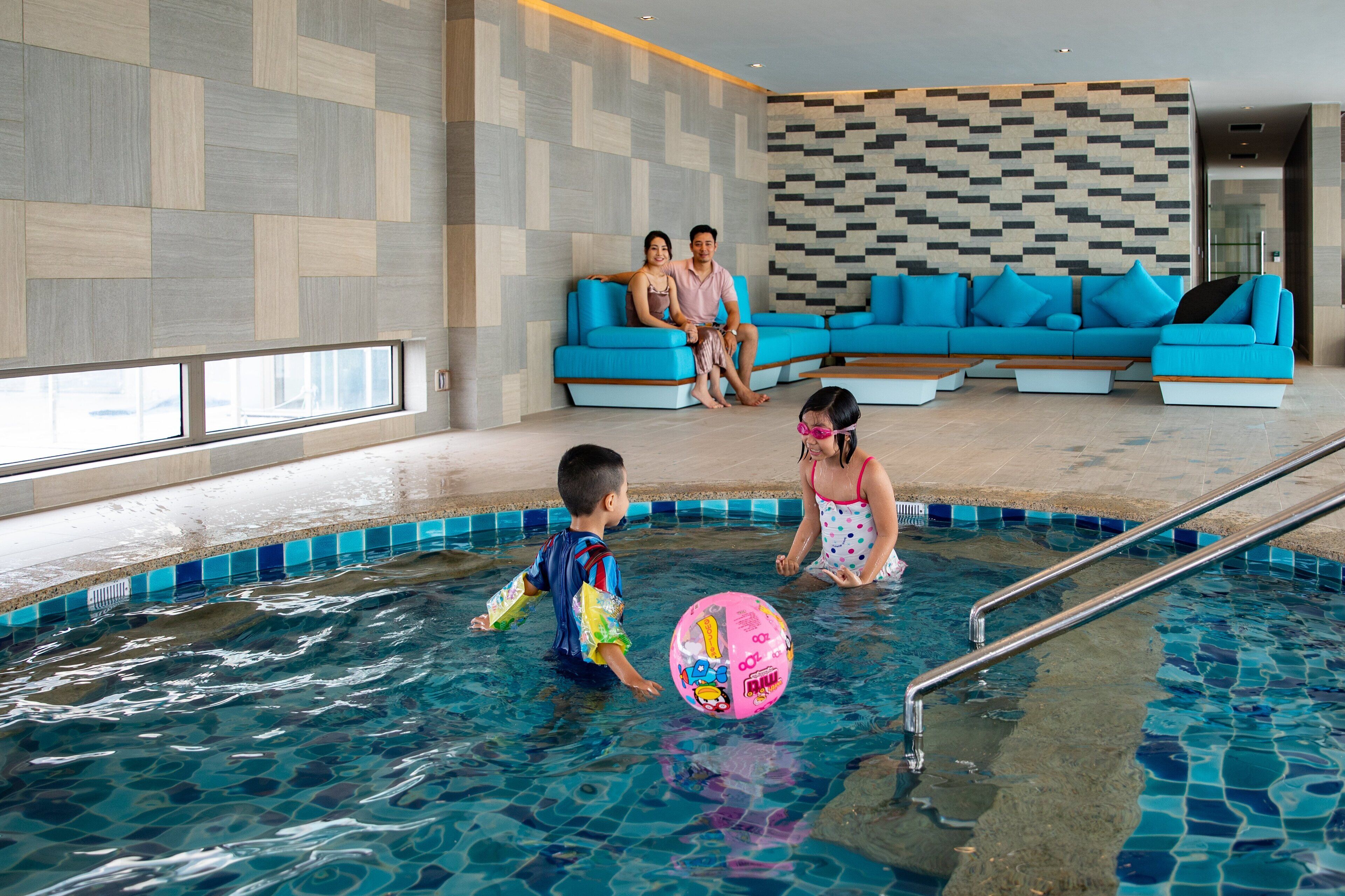 An indoor swimming pool with a blue and white checkered wall, a blue sofa, and a poolside area with a metal ladder.