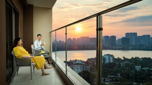 Un balcon avec vue sur la ville au coucher du soleil, deux personnes assises et une table avec une tasse.
